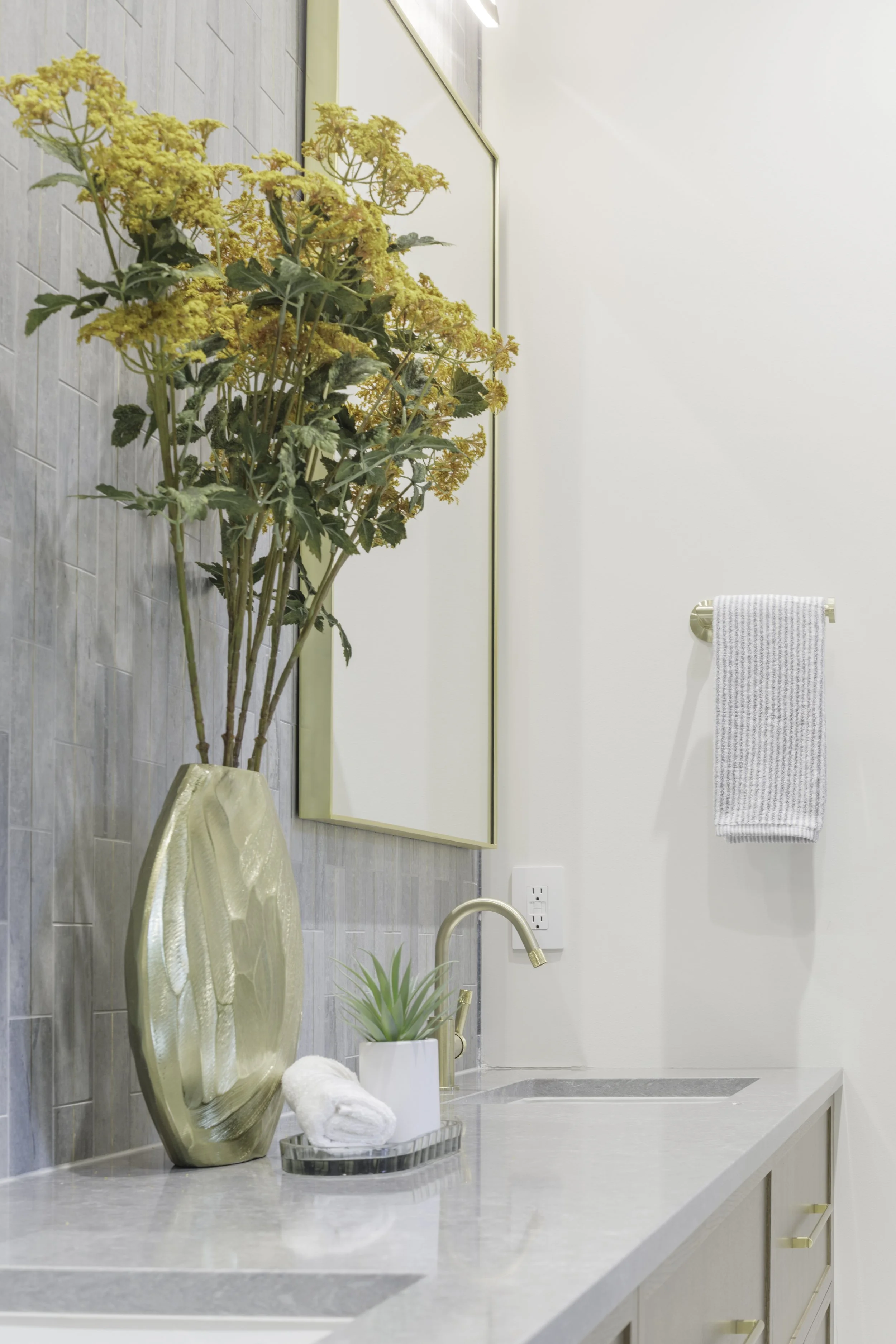Bathroom with a gold vase holding yellow flowers, a small white plant, a towel, and a gold decorative tray on a light gray countertop. A large mirror is above the counter, and a towel is hanging on a gold towel bar on the white wall.