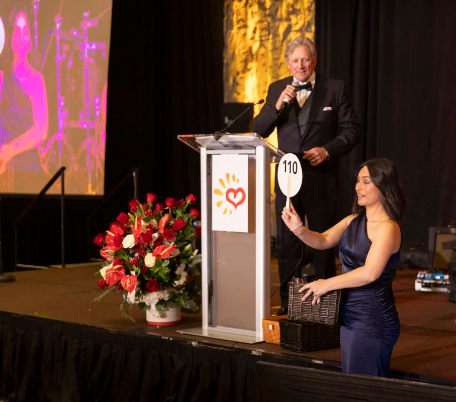 A woman in a dark blue dress, holding a sign with the number 110, is smiling and standing next to a podium with Chad Chance in a tuxedo speaking into a microphone at an event or auction.