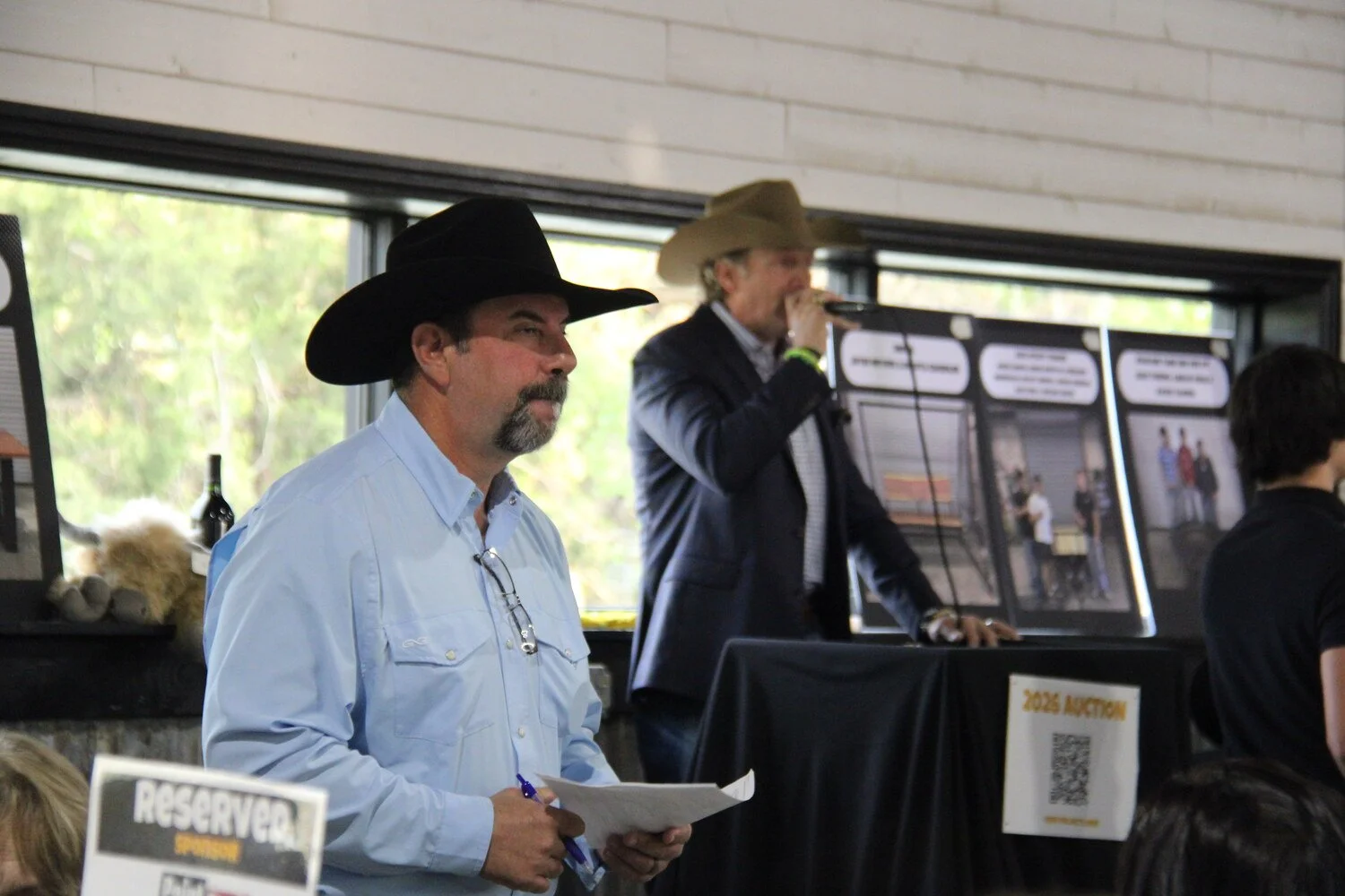 Man in a light blue shirt and cowboy hat holding notes, standing next to a reserved sign, with another man in a dark suit and cowboy hat speaking into a microphone in the background during an auction event.