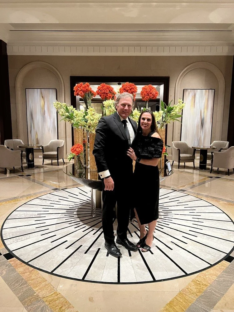 A man and woman dressed in formal attire standing together inside a hotel lobby with large floral arrangements behind them. The man is in a black tuxedo and the woman is in a black dress. They are smiling and posing for the photo on a decorative circular rug.