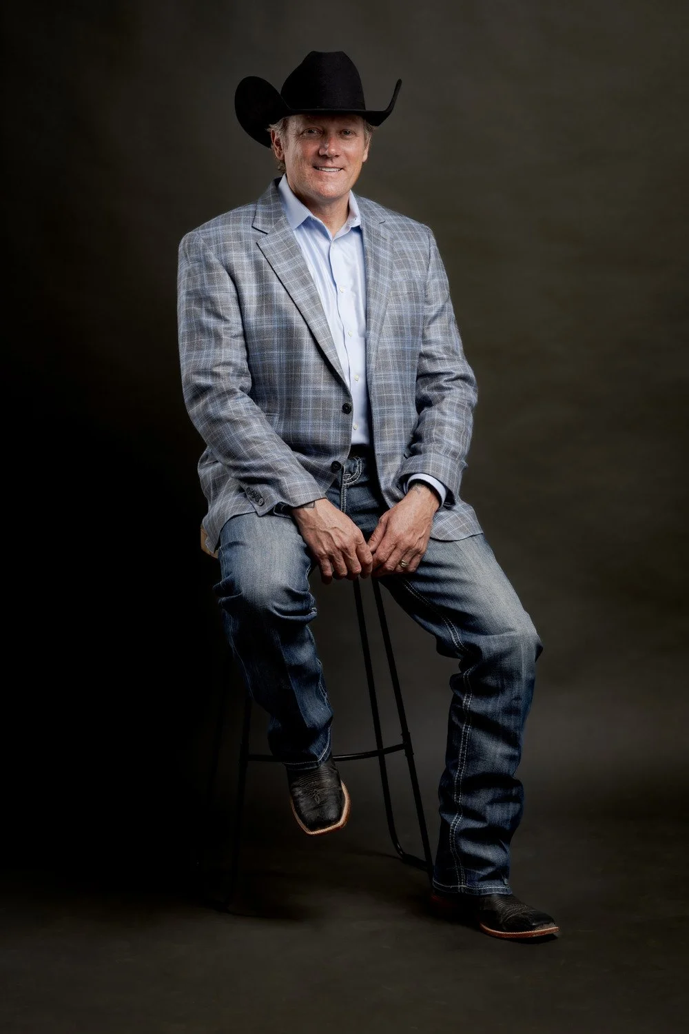 Chad Chance wearing a large black cowboy hat, a light checkered blazer, a light blue dress shirt, jeans, and cowboy boots sitting on a stool against a plain dark background.