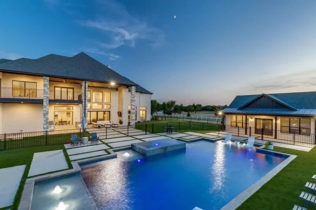 View of a luxurious backyard at dusk featuring a swimming pool, a large two-story house with balconies, and a separate pool house, surrounded by a green lawn and enclosed by a black fence.