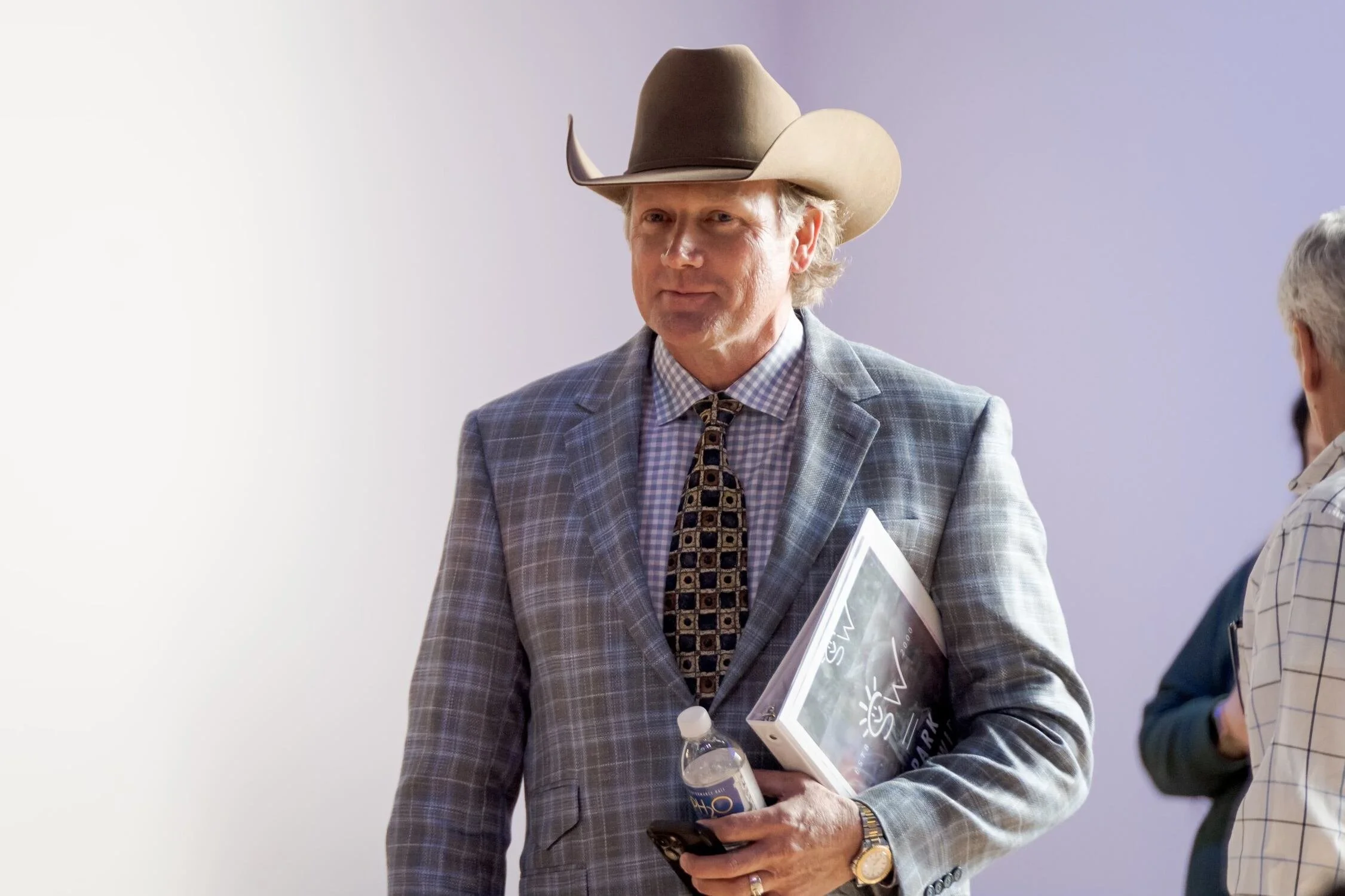 Chad Chance wearing a cowboy hat, a plaid suit jacket, and a patterned tie, holding a water bottle and a book, and standing with a woman in a striped blazer and holding a magazine,.
