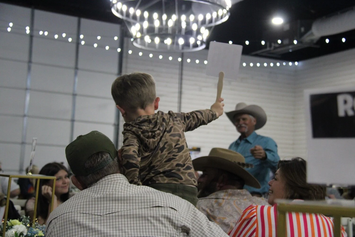 A child with short blonde hair, wearing a camouflage hoodie, is raising a wooden stick with a blank white sign attached, facing an older man with a white mustache and beard, wearing a large cowboy hat and a blue shirt, at what appears to be a social event or gathering. The setting resembles a barn or hall with string lights hanging from the ceiling, and several adults and children are visible in the background, some wearing cowboy hats.