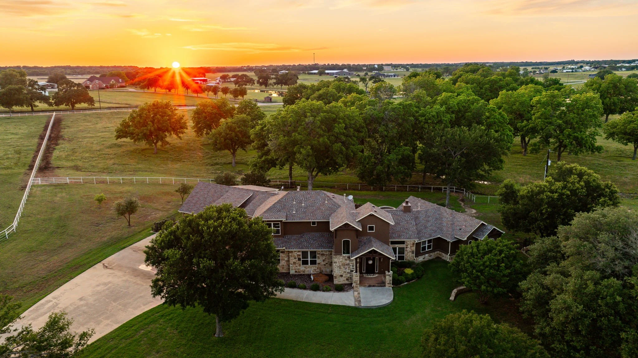 Aerial view of a rural landscape at sunset featuring a large house surrounded by trees, green fields, fences, and distant farm buildings.