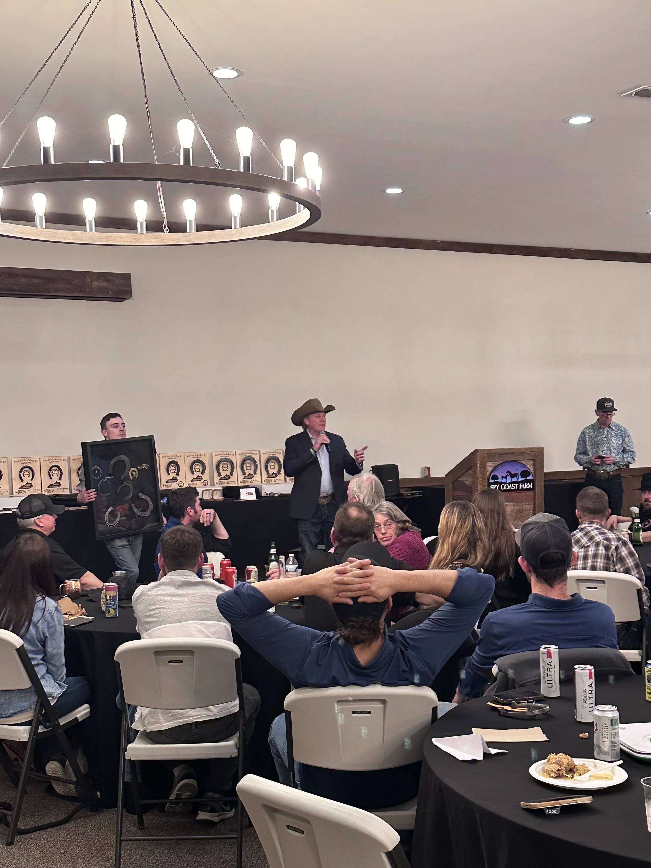A man wearing a cowboy hat and suit speaking into a microphone at an event, surrounded by an audience seated at round tables with food and drinks, and a woman with a clipboard in the background. Large chandelier hangs from the ceiling.