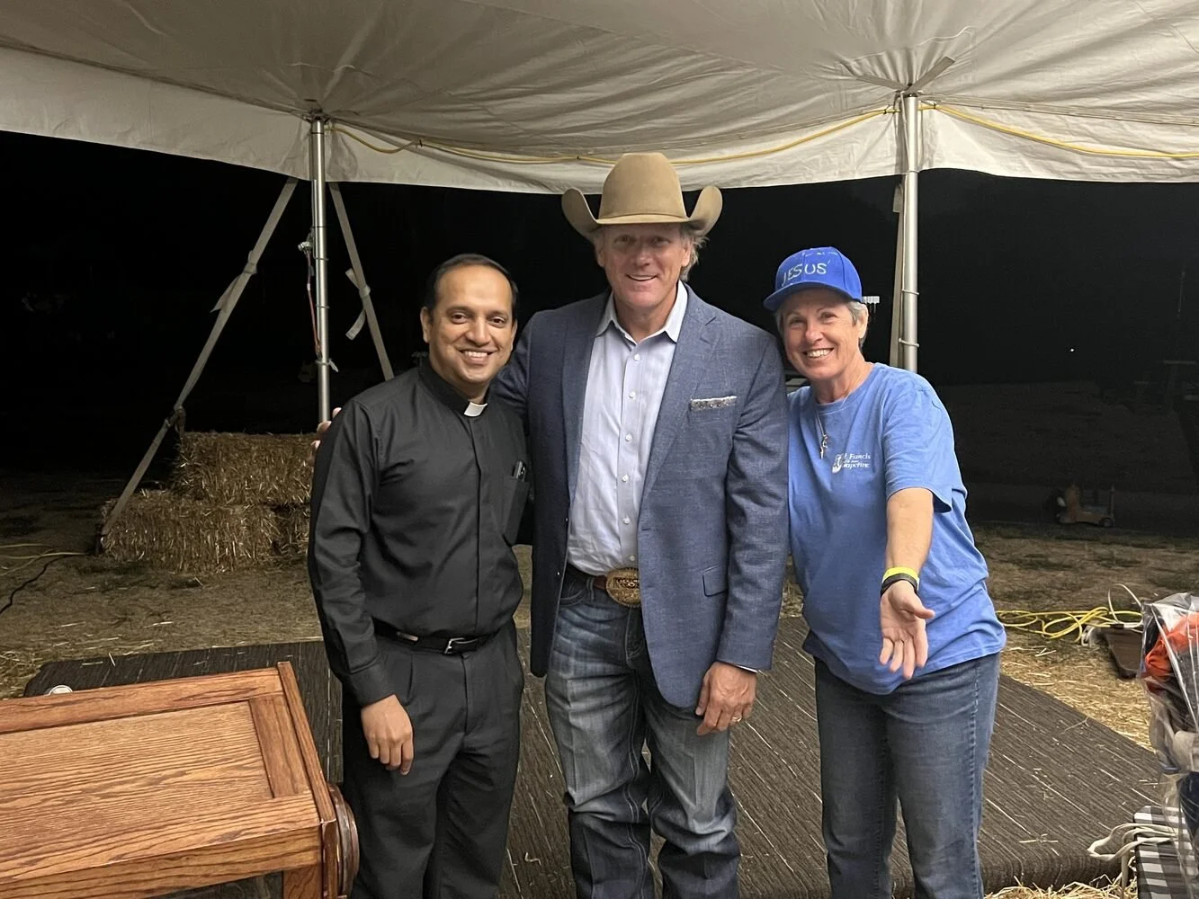 Three people standing close together inside a large tent at night. The person in the middle is wearing a cowboy hat and a suit, while the person on the right is wearing a blue shirt and a blue cap with 