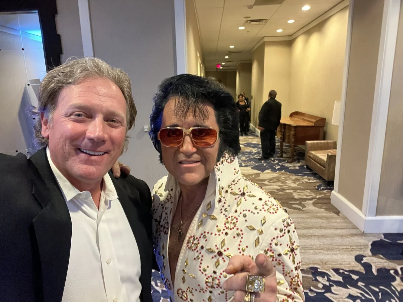 A man and a woman taking a selfie at an indoor event. The man has light skin, shoulder-length hair, and is wearing a black suit with a white shirt. The woman has tan skin, dark hair in a pompadour style, large sunglasses, an ornate white jacket with gold and red embellishments, and is wearing large rings and jewelry.