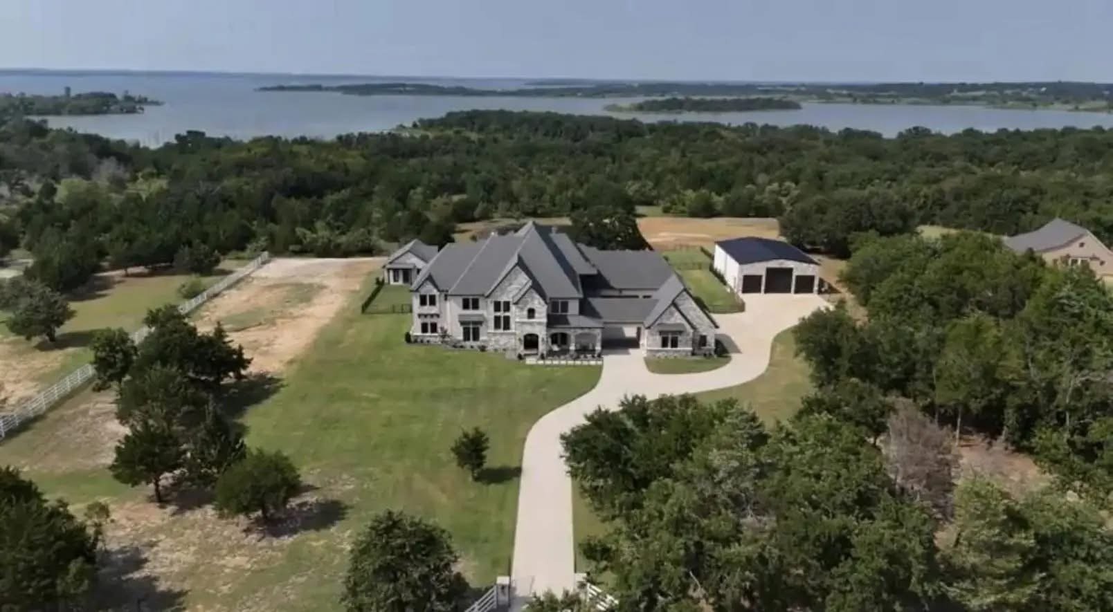 A large, gray house with a curved driveway surrounded by trees and a large lawn, with a lake in the background.