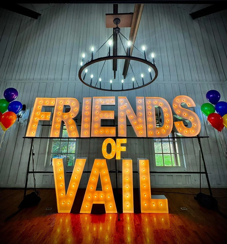 Decorative sign that says "FRIENDS OF VAIL" with large illuminated letters, surrounded by colorful balloons, inside a room with a wooden floor and white wooden walls.