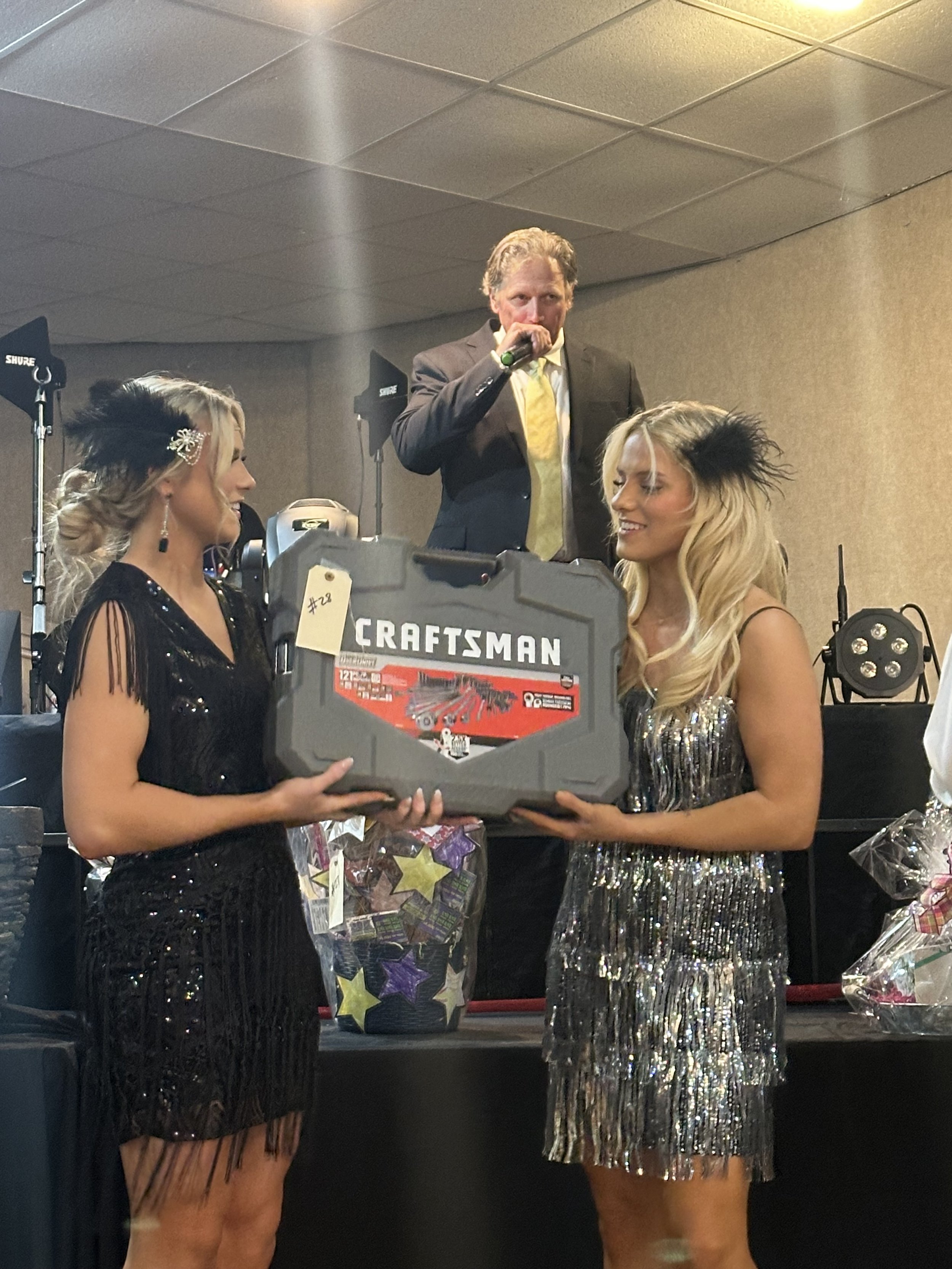 Two women dressed in black and silver dresses are accepting a large toolbox as a gift at an indoor event, with a man in a dark suit and yellow tie speaking into a microphone in the background.