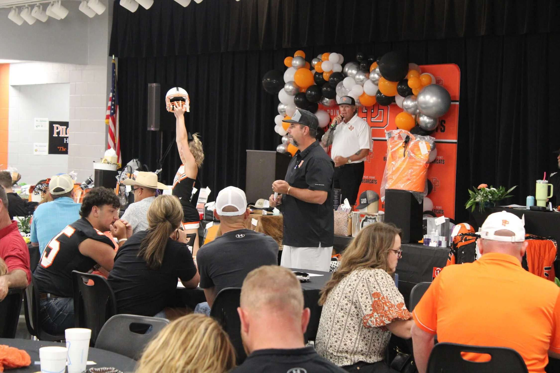 Crowd of people attending an indoor event with a stage decorated with black, white, orange, and silver balloons, and a man speaking into a microphone. Participants are wearing sports jerseys, some in black and orange, with a woman raising a small football helmet in the air.