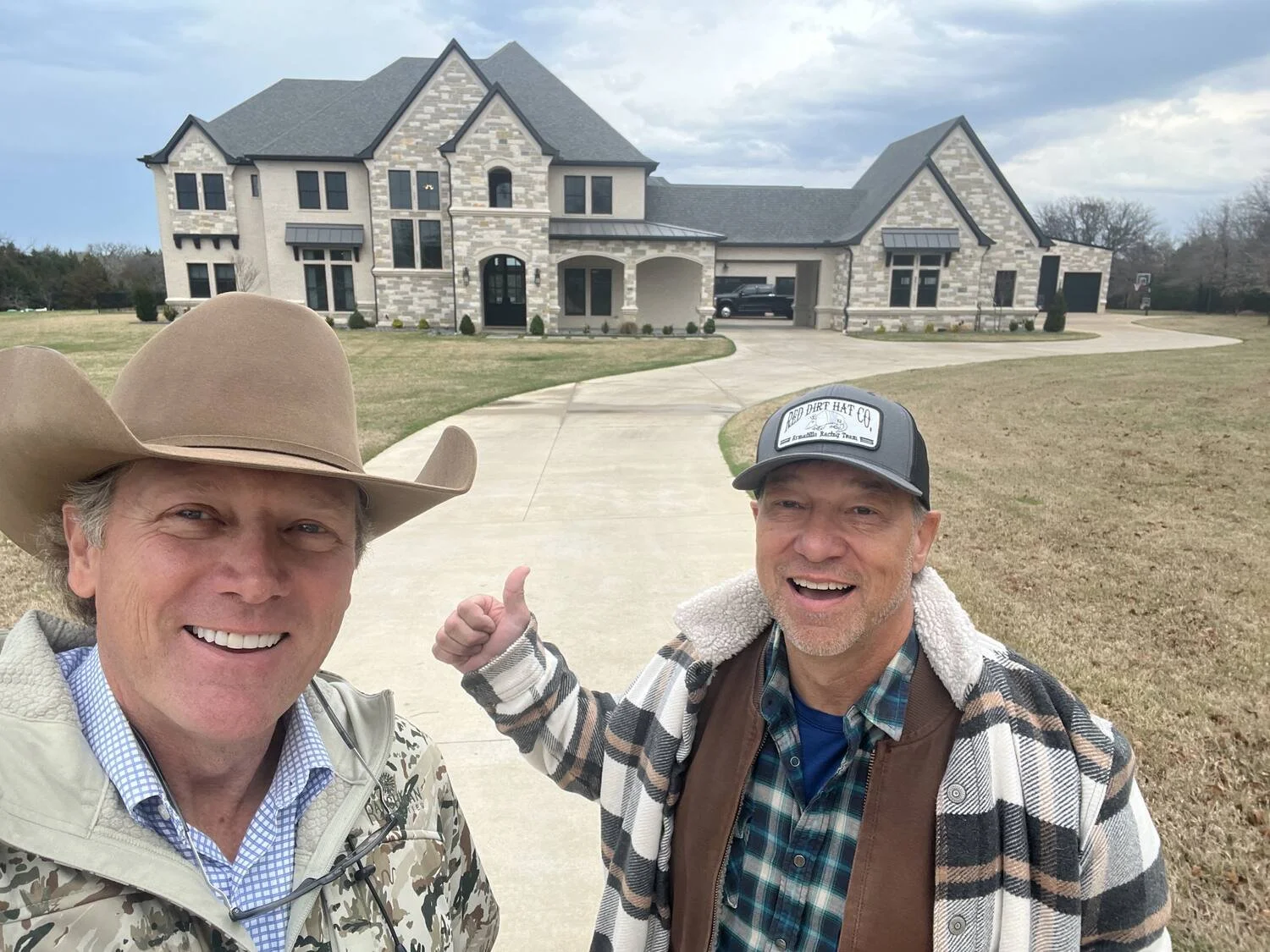 Two men smiling in front of a large, modern house with a driveway, one wearing a cowboy hat, and the other wearing a baseball cap and plaid shirt, pointing at the house.