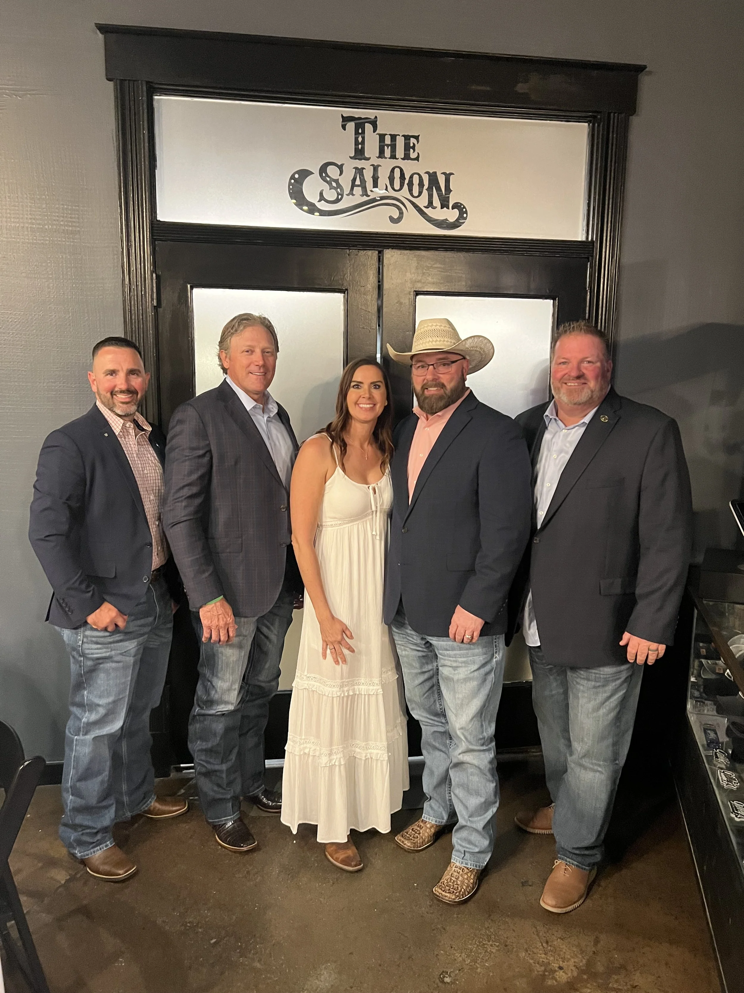 Group of five people standing in front of a sign that reads 'The Saloon'. Four men and one woman, dressed in business casual and western-style attire, smiling at the camera.