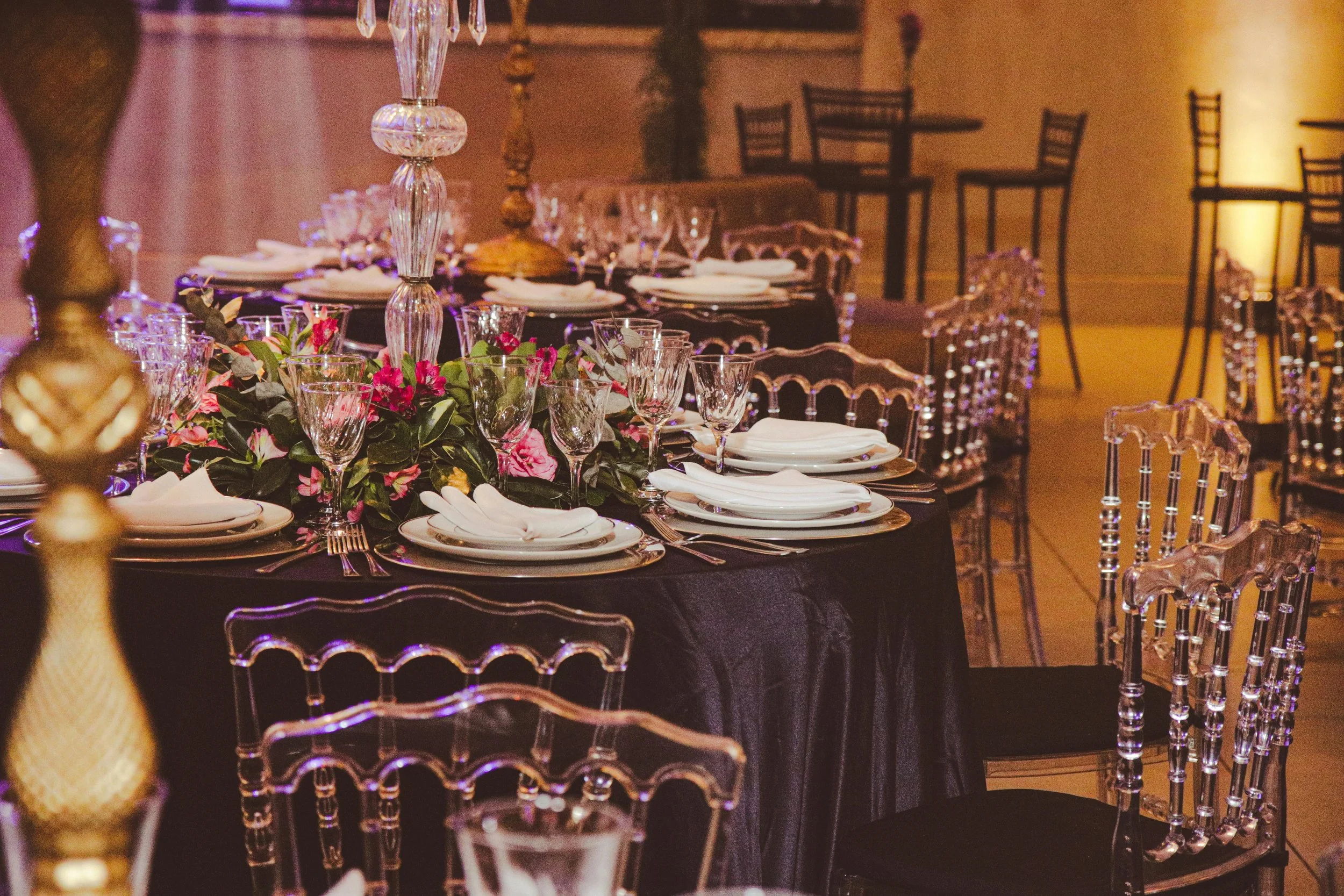 A banquet table set for an event with elegant glassware, white plates, folded napkins, and a floral centerpiece with pink flowers and greenery. Transparent chairs surround the table, and chandeliers and other tables are visible in the background.