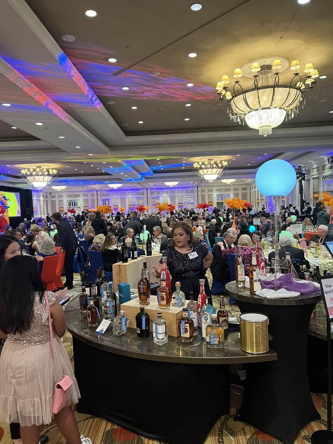 A large banquet hall filled with numerous guests seated at decorated tables. There are multiple chandeliers and colorful lighting, with balloons and decorative flowers. A booth displays bottles of alcohol.