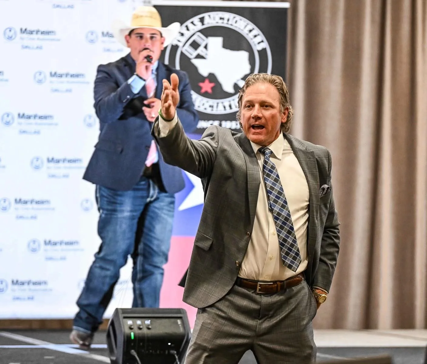 Two men in a professional setting, one speaking passionately and gesturing with his hand, the other standing behind him, holding a microphone, with a backdrop featuring the Texas flag and logos.