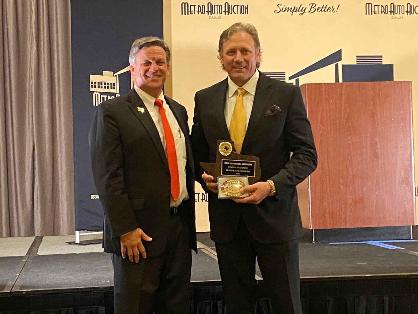 Two men in business suits at an award ceremony, one holding a plaque and medal. Background features a yellow banner with black and white logos and text, with a gray curtain on the left.