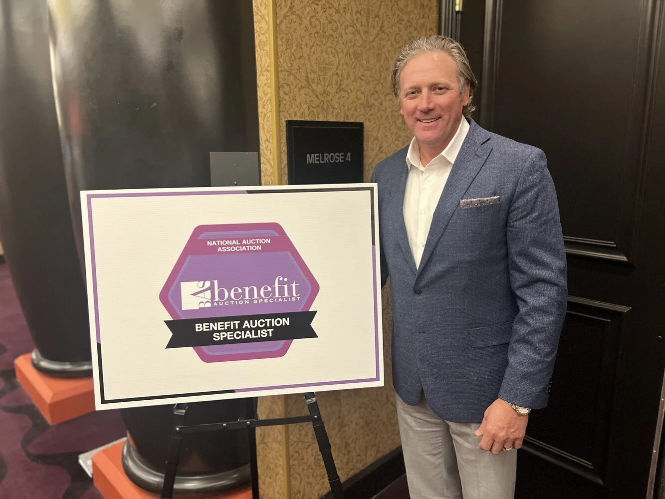 Chad Chance in a blue blazer and white shirt standing next to a sign that says Benefit Auction Specialist, at a benefit auction event.