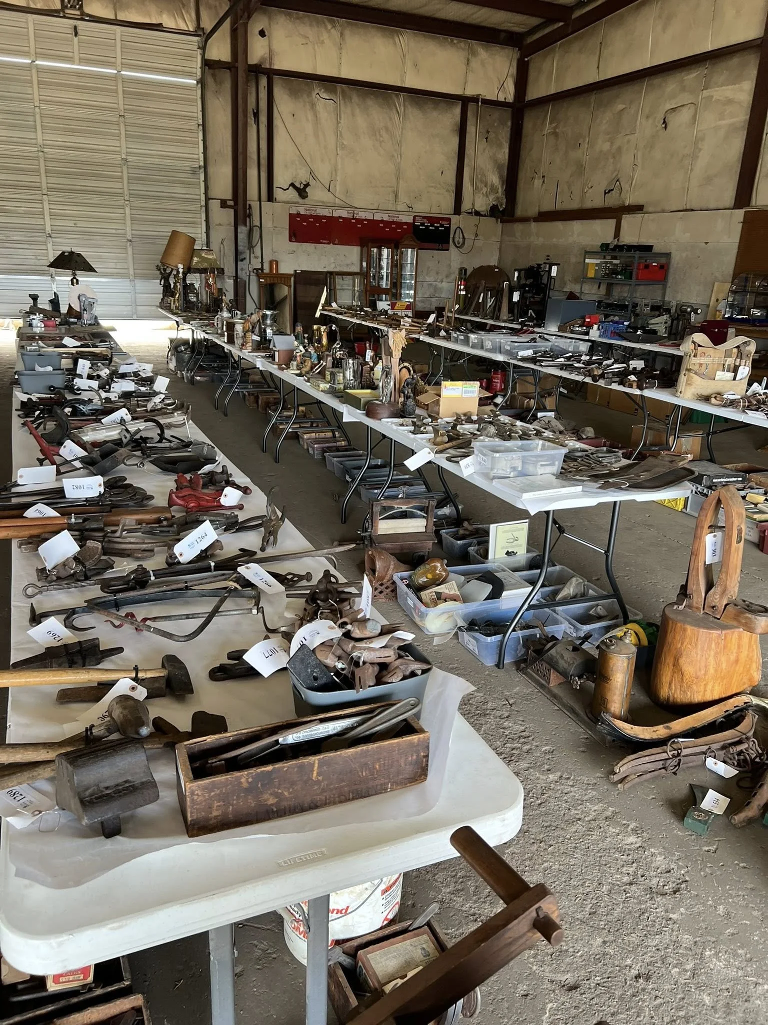 Tables filled with vintage tools and relics in a warehouse or auction setting, with handmade tags and some furniture pieces visible.