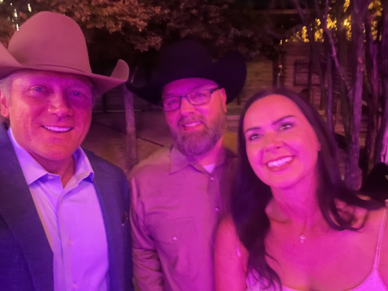 Three people smiling at a night event, wearing cowboy hats and casual attire, with trees and a fence in the background.