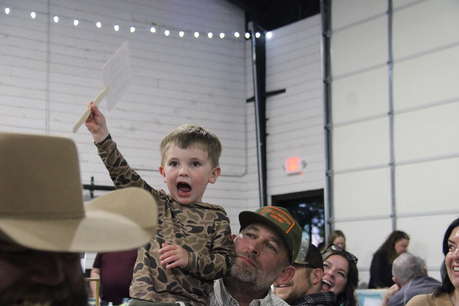 A young boy wearing camouflage shirt holding a paper and what appears to be a shoehorn or similar object, sitting on a man's shoulders during an indoor event with other people smiling and watching.