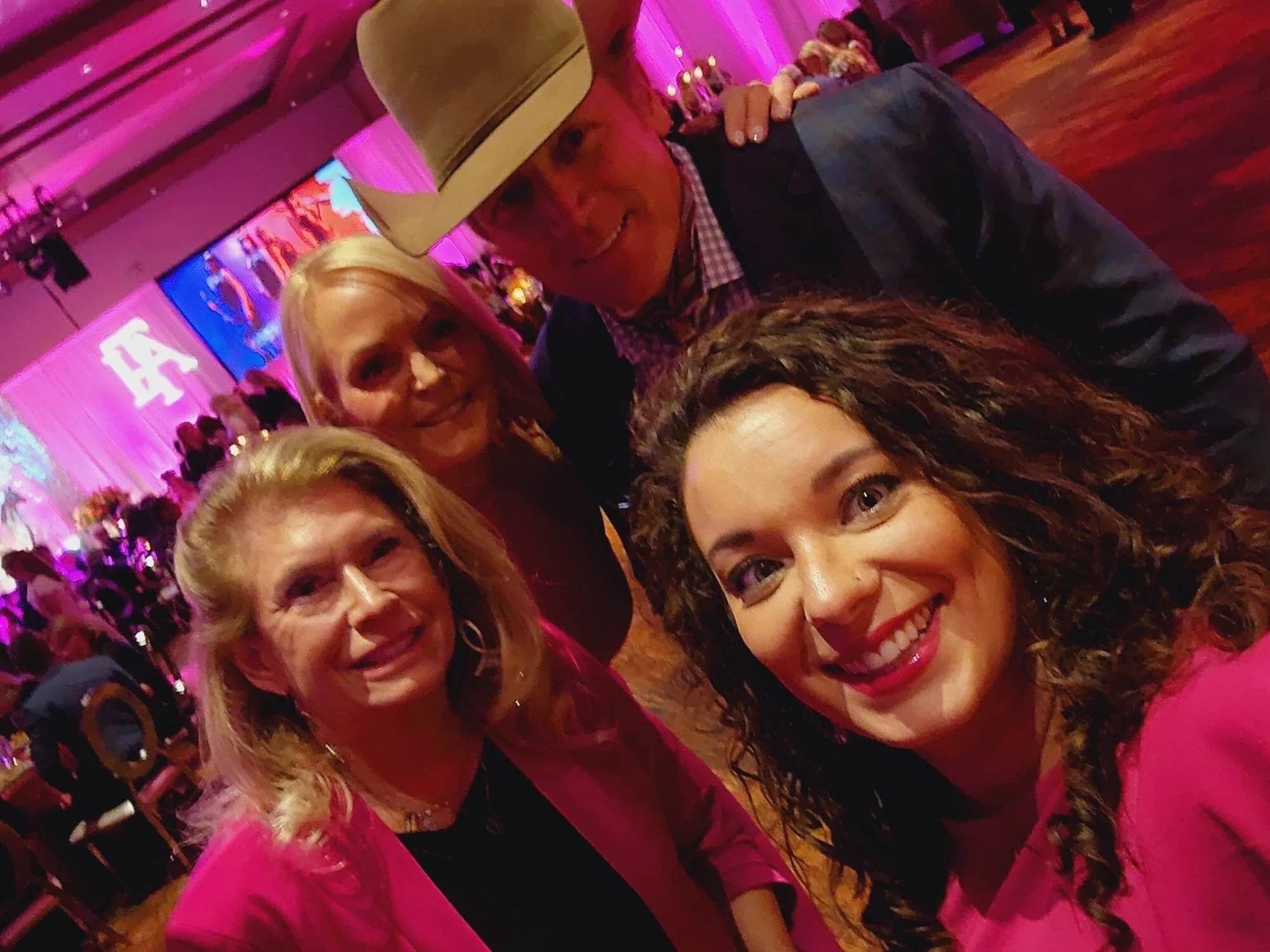 A group of women at a celebration, with a woman wearing a cowboy hat in focus, smiling, with pink and purple lighting in the background.
