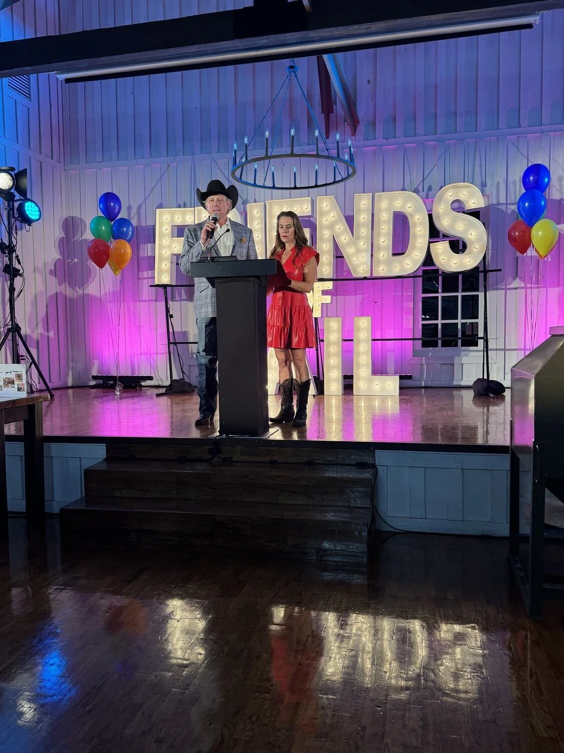 Two people on a stage with a large illuminated 'FRIENDS' sign behind them, decorated with balloons. The man on the left is wearing a cowboy hat and blazer, holding a microphone. The woman on the right is in a red dress and black boots, looking at the man. The stage has lighting with pink and purple hues.