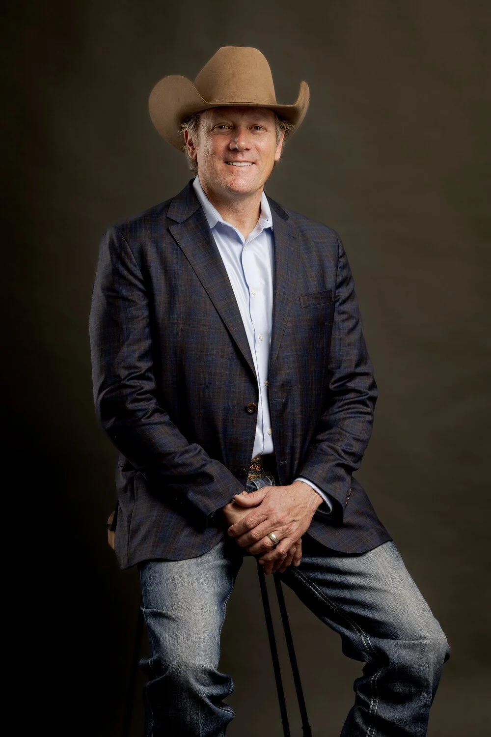 Chad Chance wearing a cowboy hat, a blue plaid blazer, and jeans smiling at the camera against a brown background.