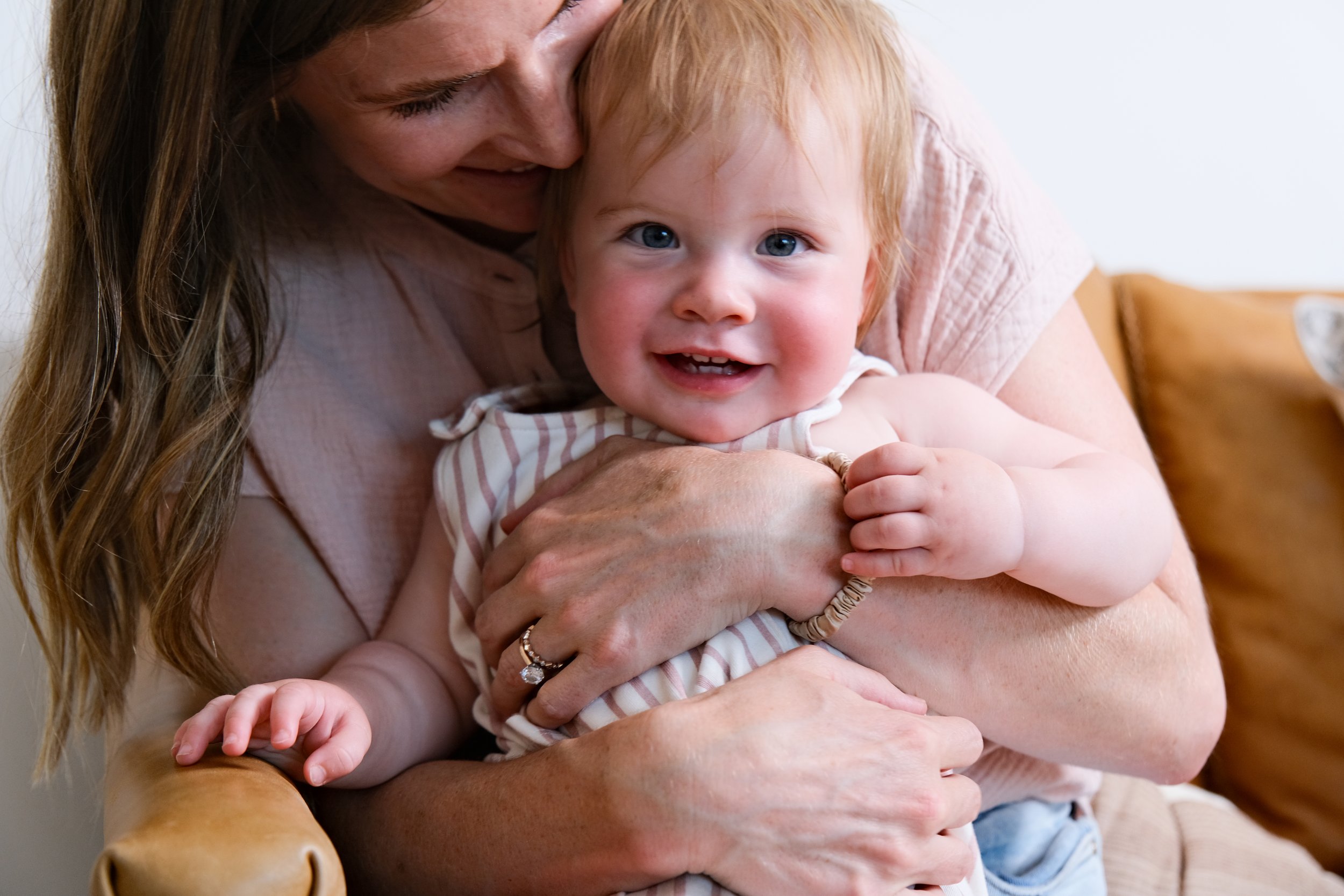 A mother happily holding her smiling baby in her arms while sitting on a leather couch.