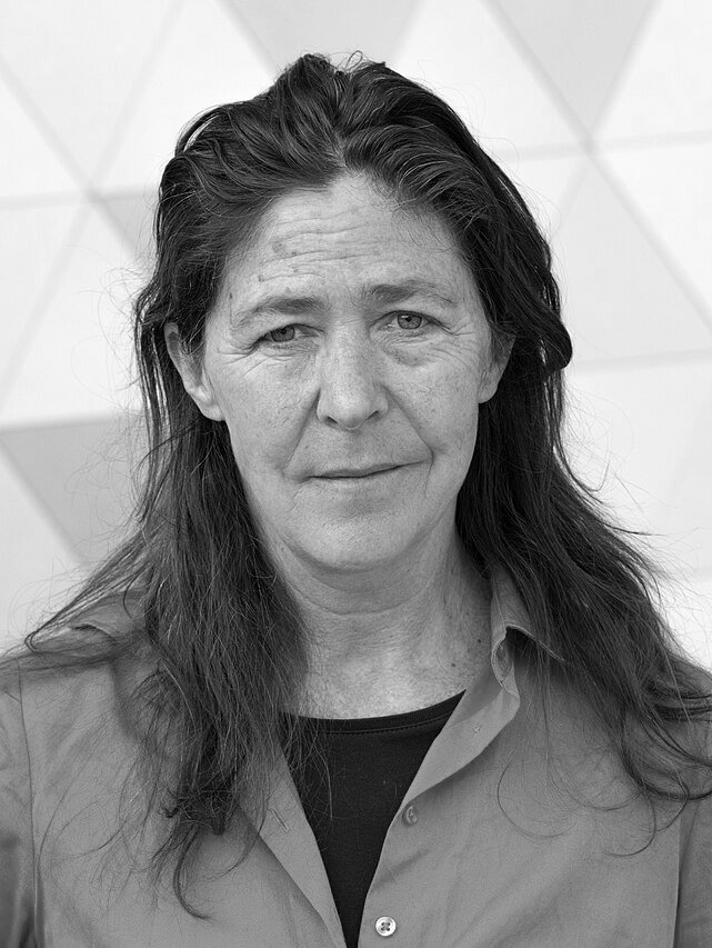 Schwarz-Weiß-Porträt einer ernst aussehenden Frau mit langen, dunklen Haaren vor einem geometrischen Hintergrund.
