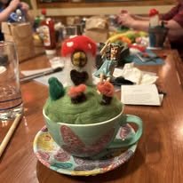 A teacup filled with a green cupcake decorated to look like a dinosaur with red and black eyes, a red crest, and tiny green arms, surrounded by a colorful plate on a cluttered table.