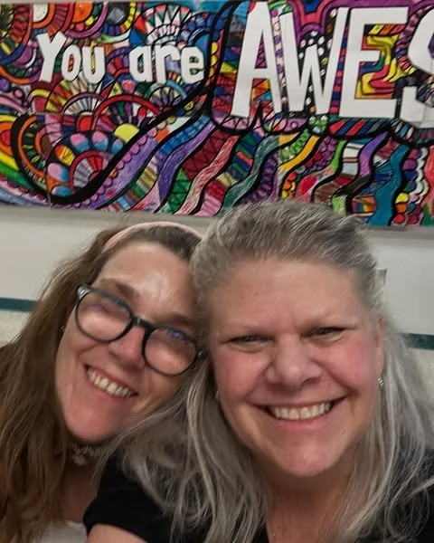 Two women smiling for a selfie in front of a colorful, abstract mural that says 'You are Awesome'.
