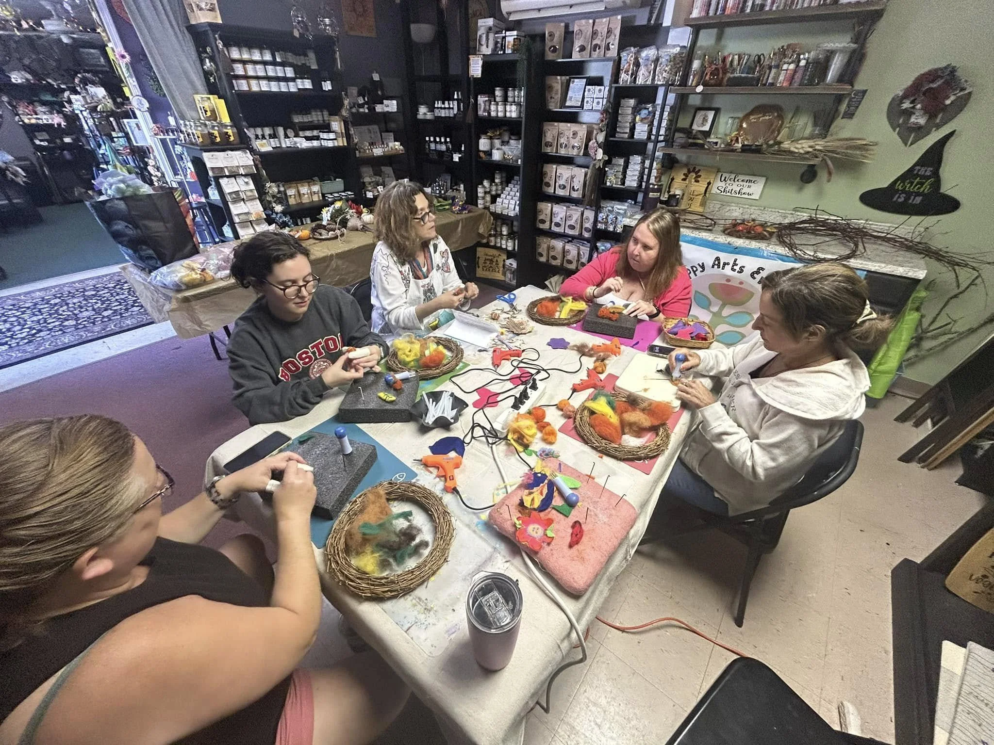 Group of women participating in a craft workshop around a table with craft supplies, felt, and glue guns in a store or craft room.