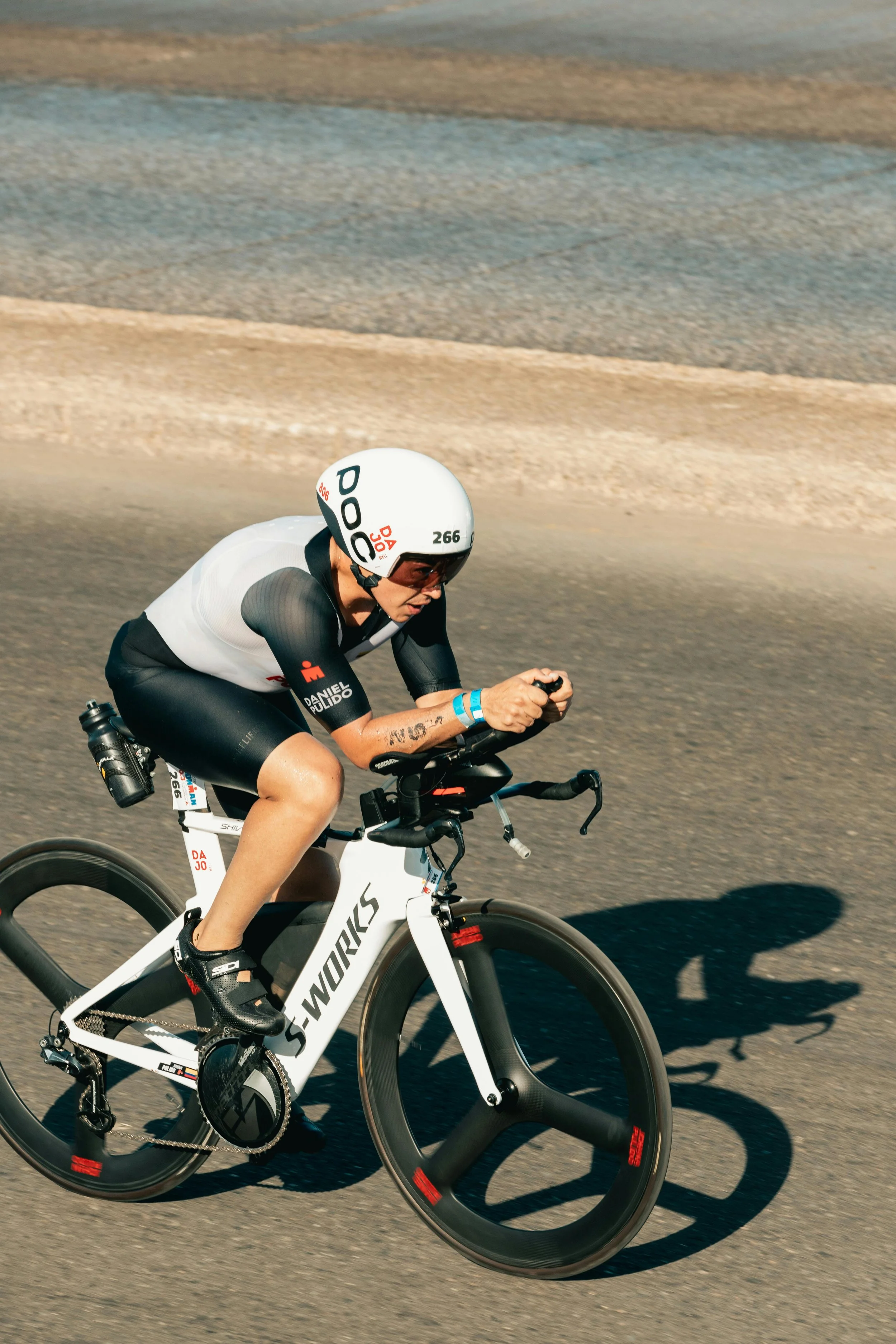 A cyclist wearing a white helmet, black and white cycling gear, riding a white S-Works bike with disc wheels on a paved road, with a blurred background.