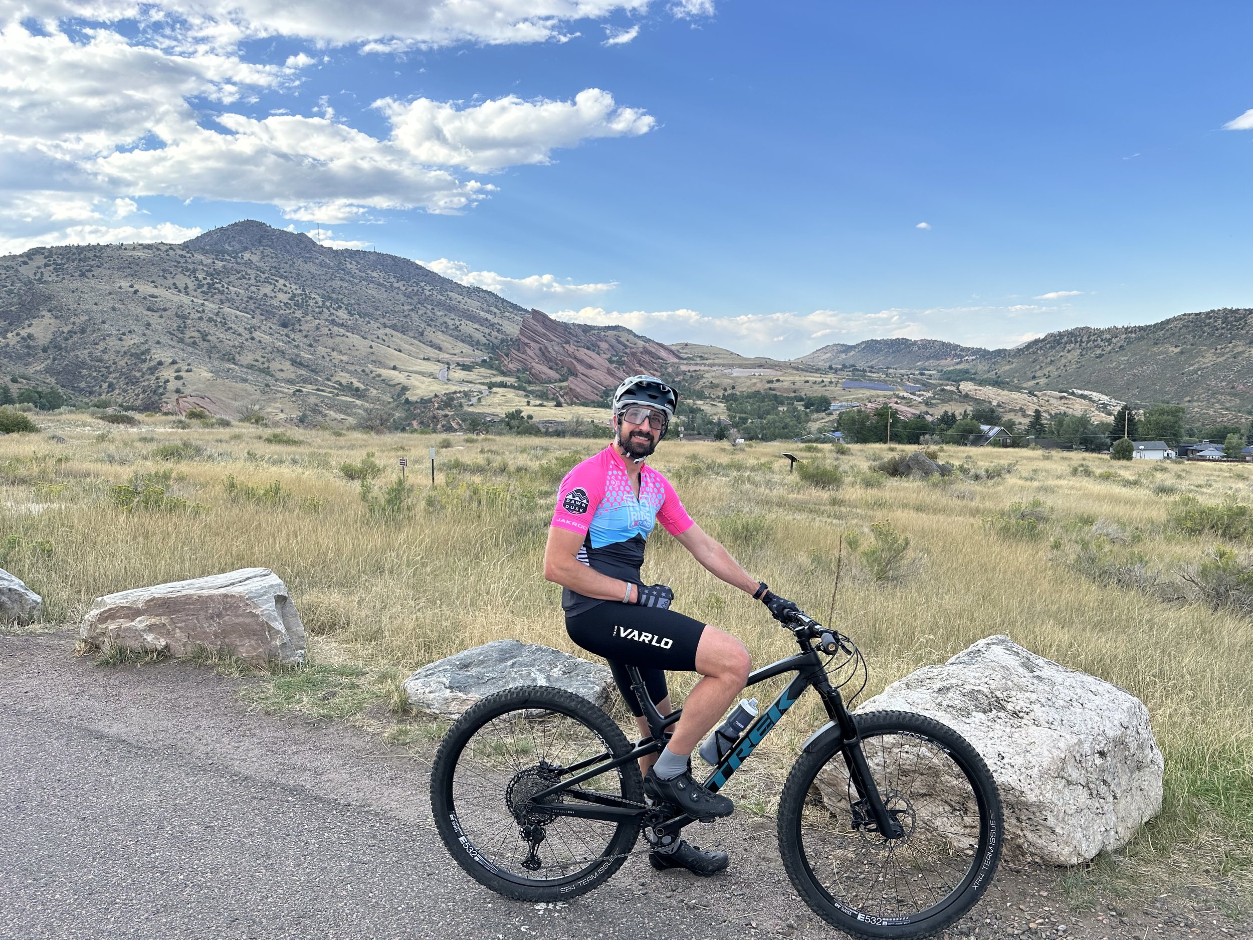 A man wearing a pink and blue cycling jersey, black shorts, and a helmet, sitting on a black mountain bike, on a dirt path with rocks and a grassy field, with mountains and a partly cloudy sky in the background.