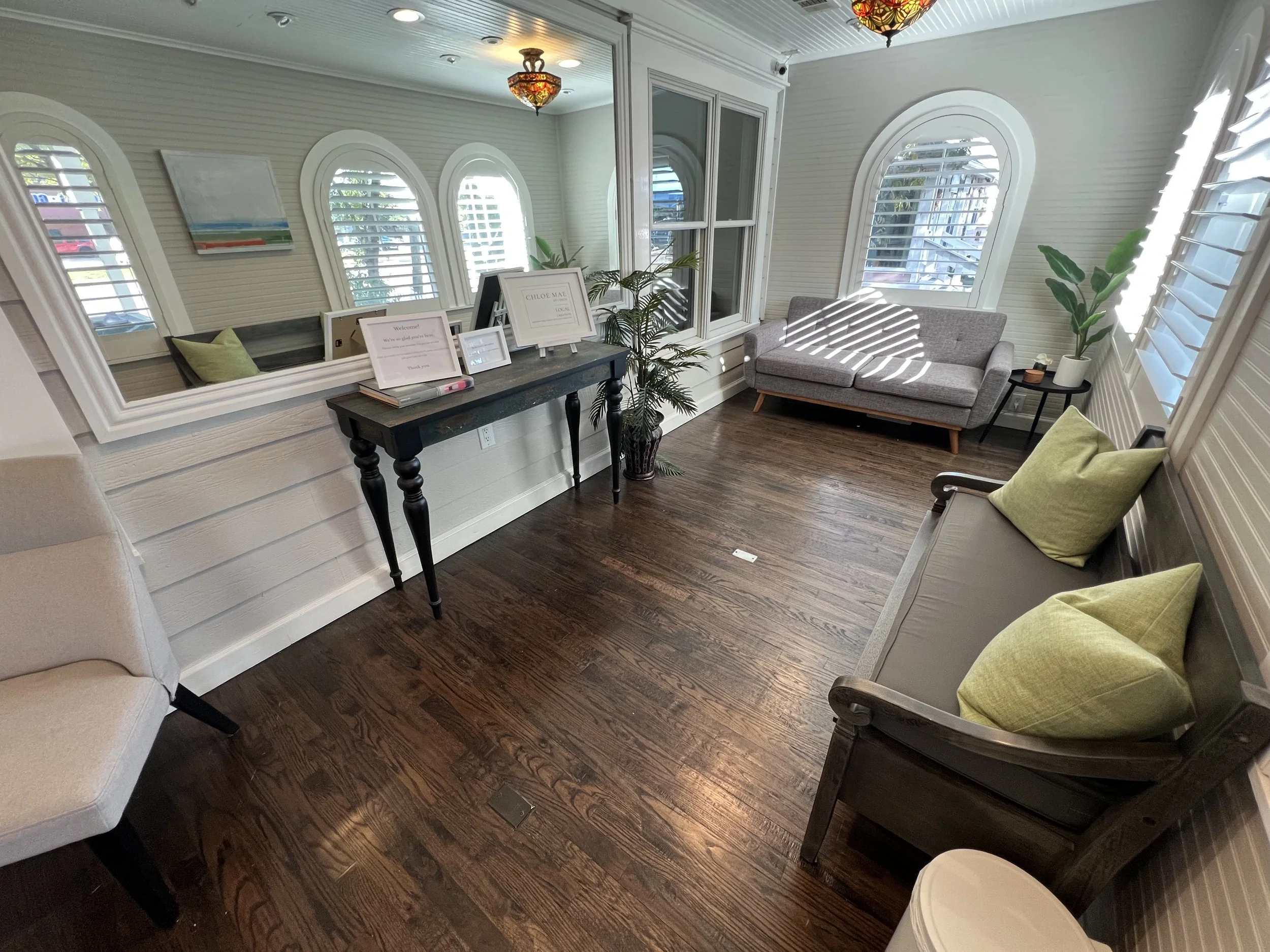 Healing House's lobby looks like a cozy living room with wooden floors, multiple windows with shutters, a mirror on the wall, a gray sofa, a bench with green pillows, a potted plant, and a black table with frames on top.