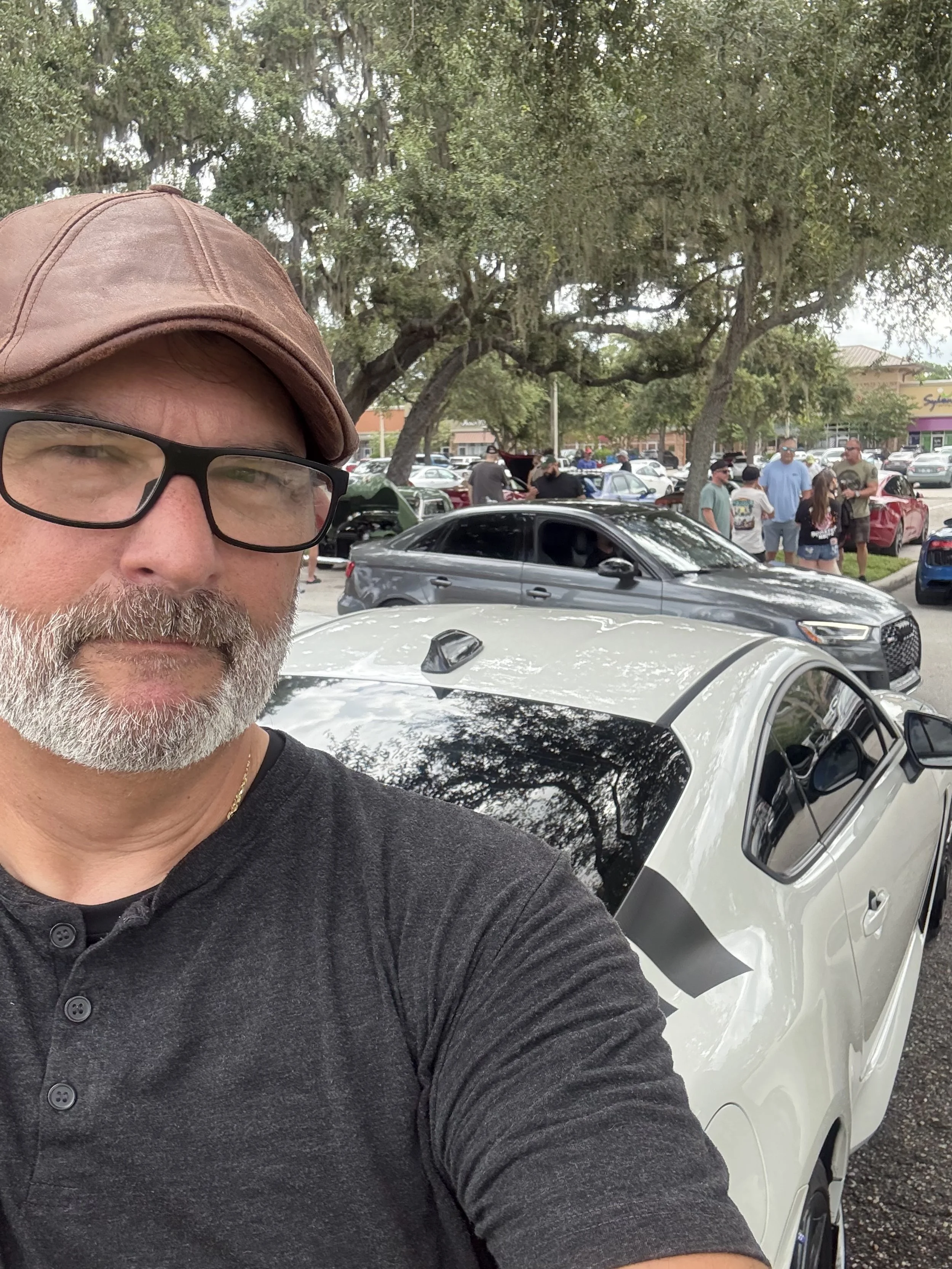 A man with glasses, a brown cap, and a gray beard taking a selfie in front of a white sports car at a car meet with other cars and people in the background under large trees.