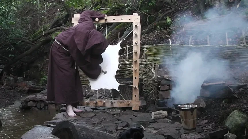 A person in a dark hooded robe making a large splash using a white liquid substance on a wooden frame outdoors near water, with smoke and a bucket nearby.
