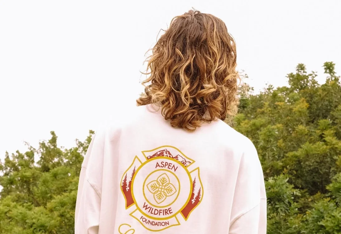 Back of a woman with curly red hair standing outdoors, wearing a white jacket with a logo for the Aspen Fire Foundation.