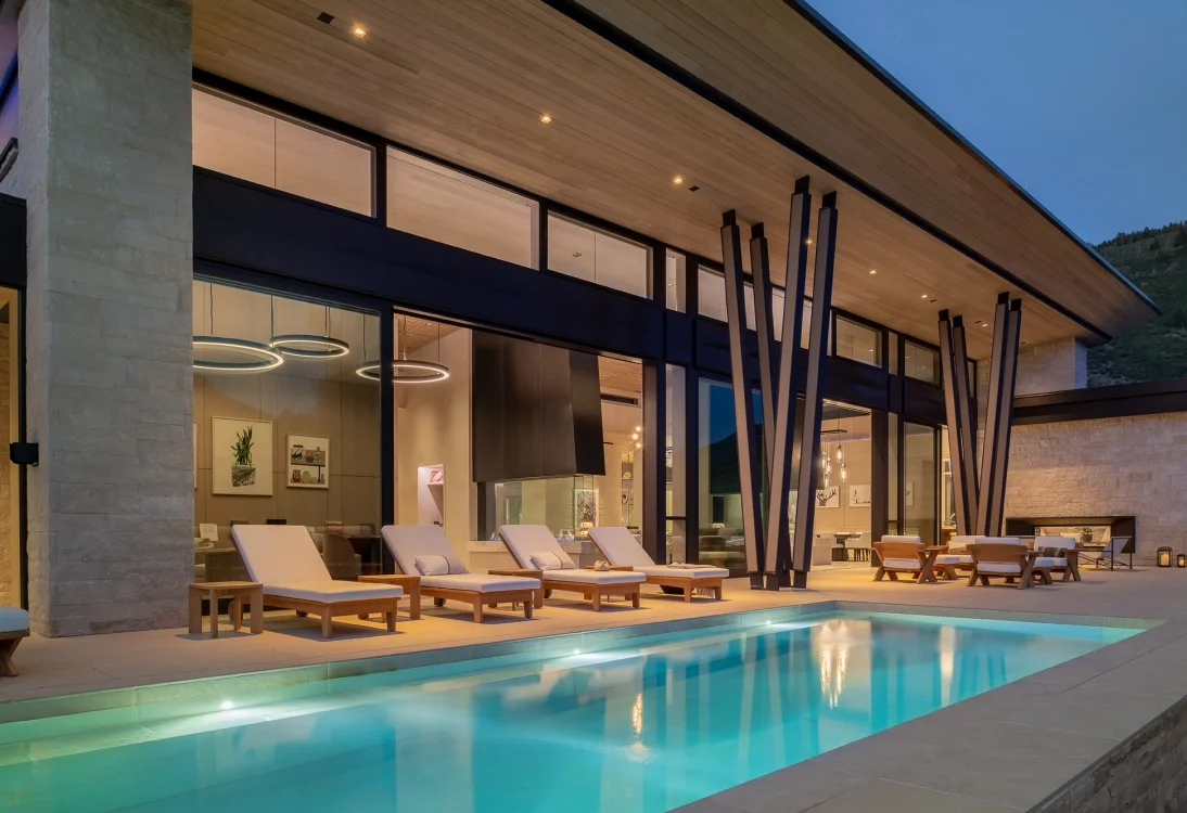 Modern house with large windows, an outdoor pool, and poolside lounge chairs at dusk.