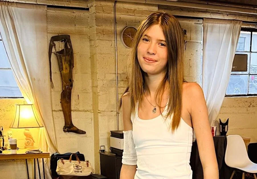A young woman with long brown hair wearing a white sleeveless top, standing in a room with beige brick walls and a window with white curtains.