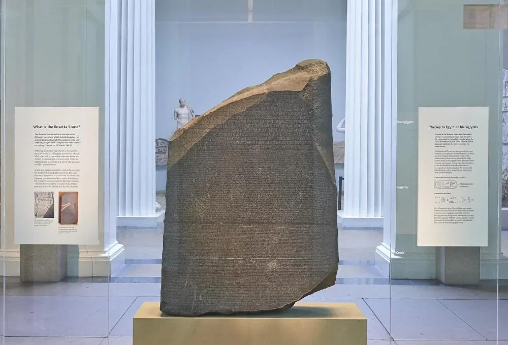 The Rosetta Stone, an ancient stone inscribed with hieroglyphics, Greek, and Demotic scripts, displayed in a museum case with informational plaques on either side.