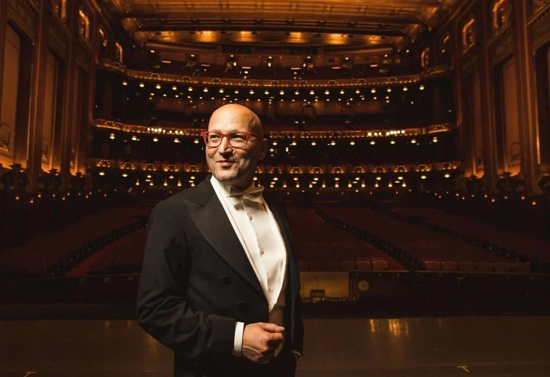 A man in a tuxedo with red glasses standing on a stage in an ornate theater with empty seats and balconies illuminated in warm lighting.