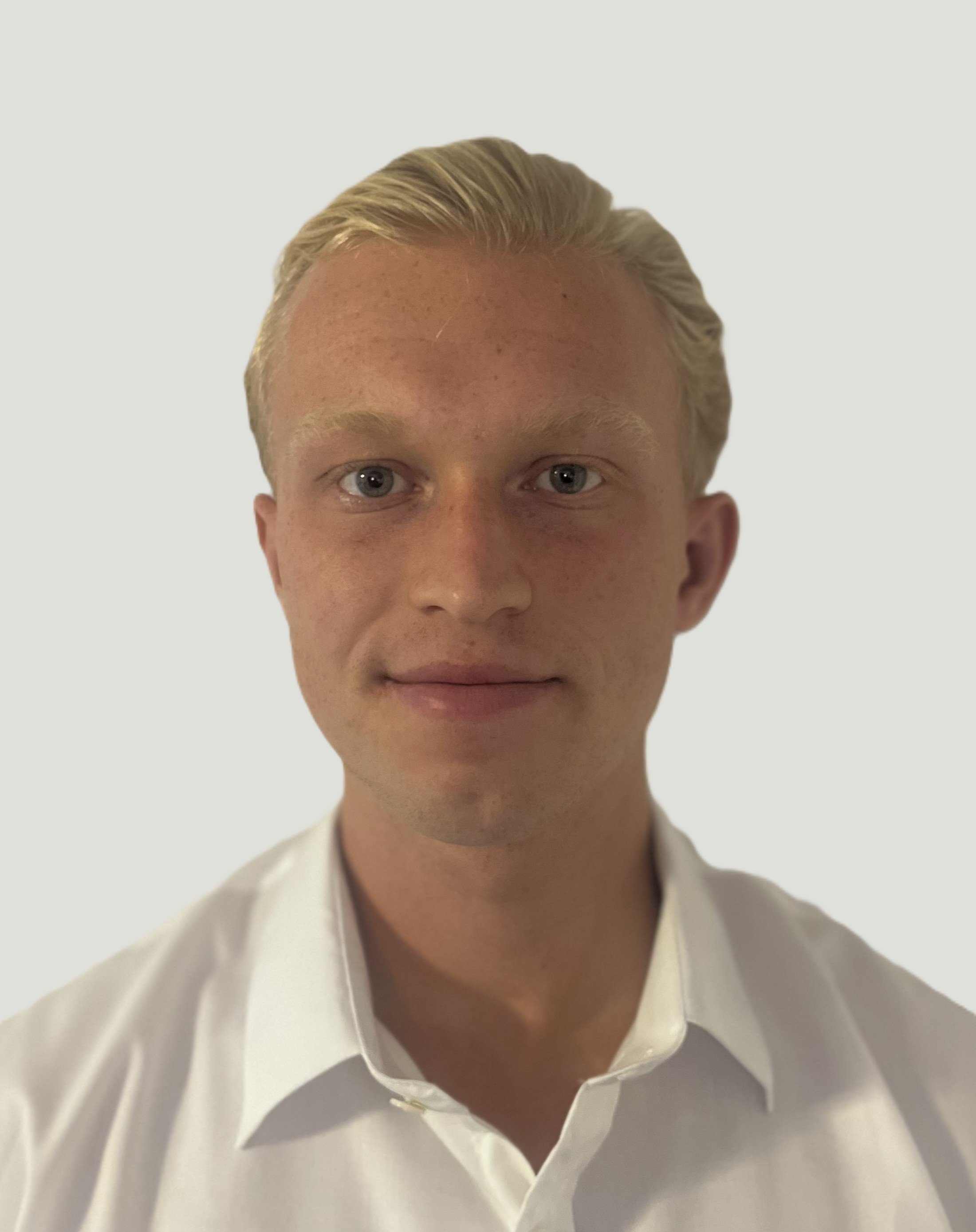 Portret van een jonge man met blond haar, blauwe ogen, gekleed in een wit overhemd, lachend, tegen een effen lichte achtergrond.