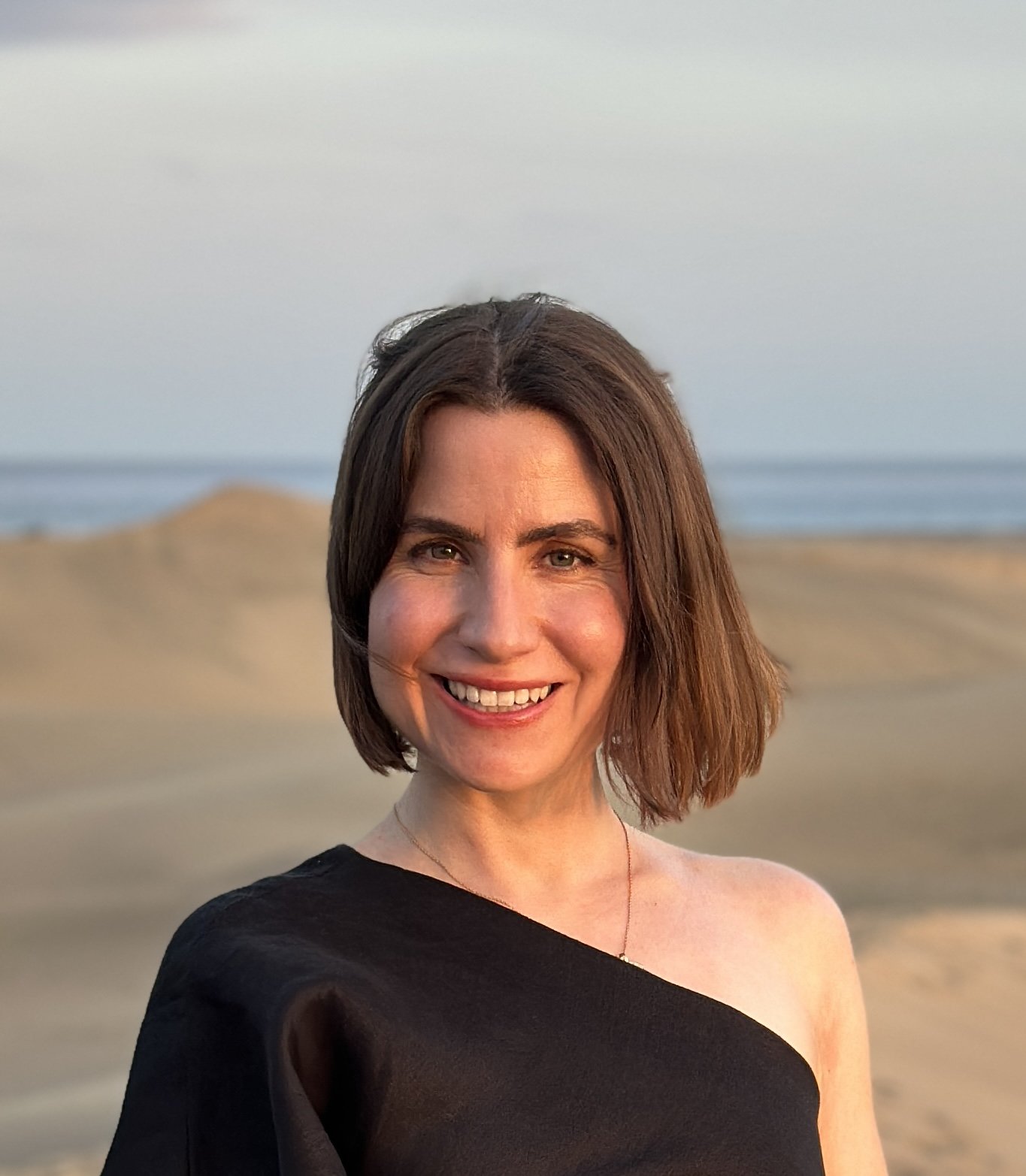 Eine lächelnde Frau mit braunen, kinnlangen Haaren trägt ein schwarzes, einseitiges Oberteil und steht am Strand bei Sonnenuntergang, im Hintergrund Sanddünen und das Meer.