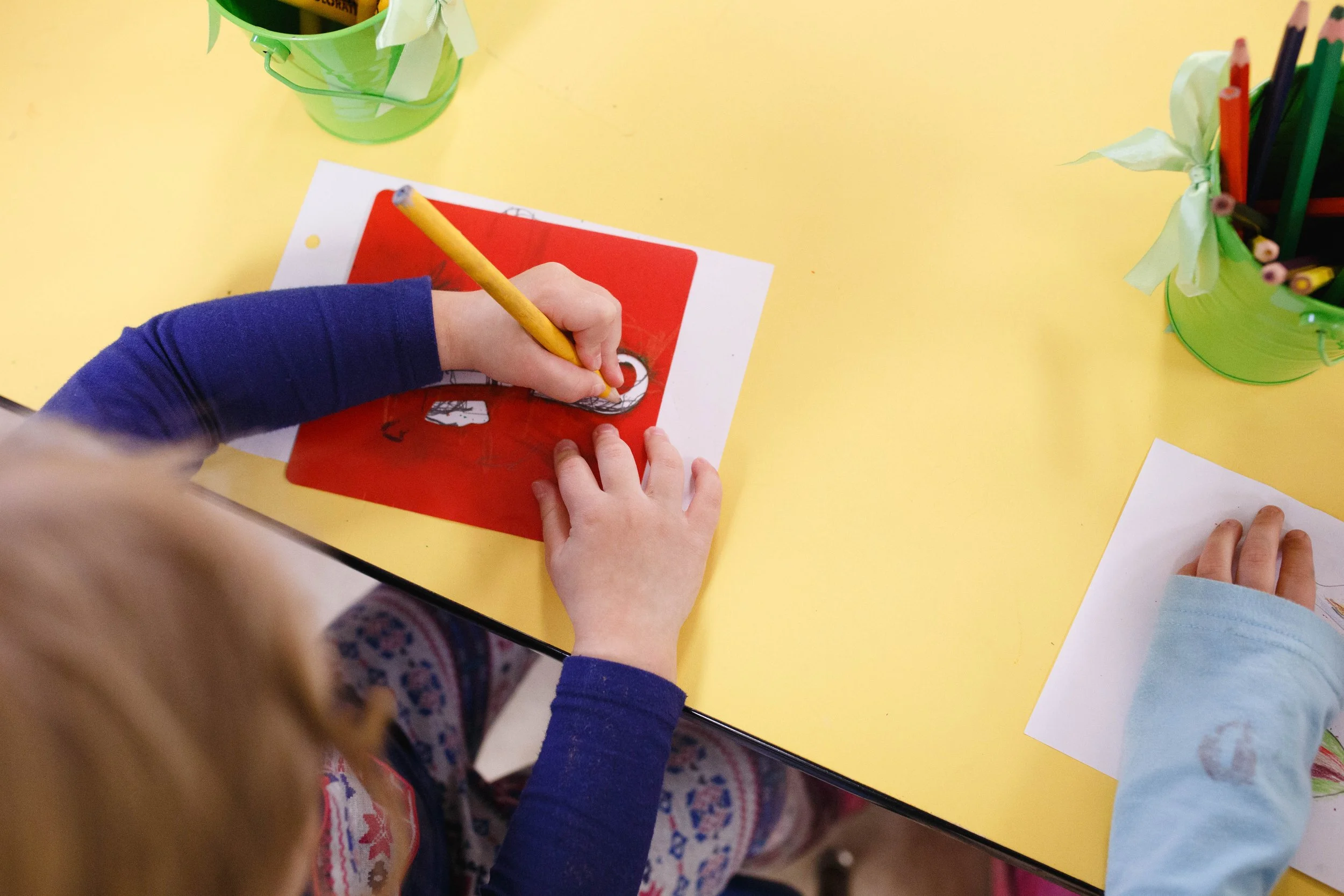 A child with a blue sleeve is coloring on a white paper with a red background using a yellow pencil at a yellow table. Next to the child is another person with a light blue sleeve. On the table are green containers holding various colored pencils.