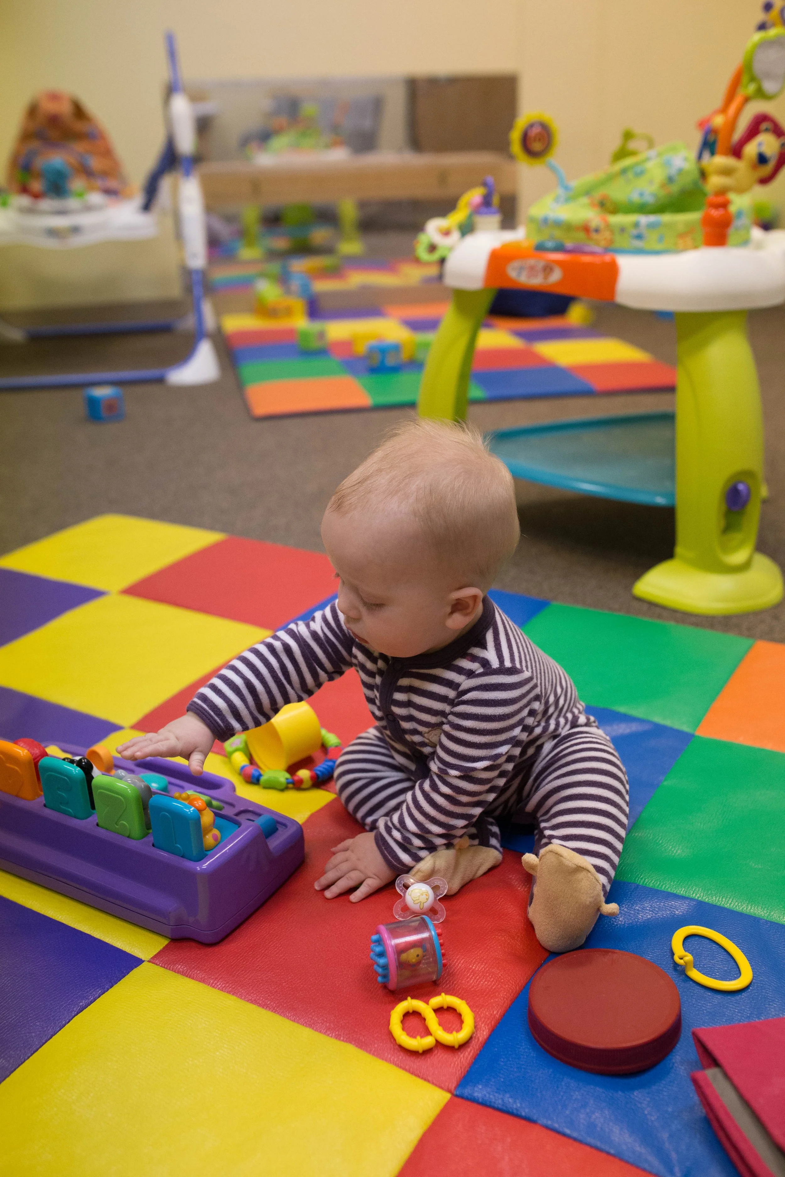 A baby sitting on colorful foam tiles playing with a purple toy that has numbered and shaped buttons.