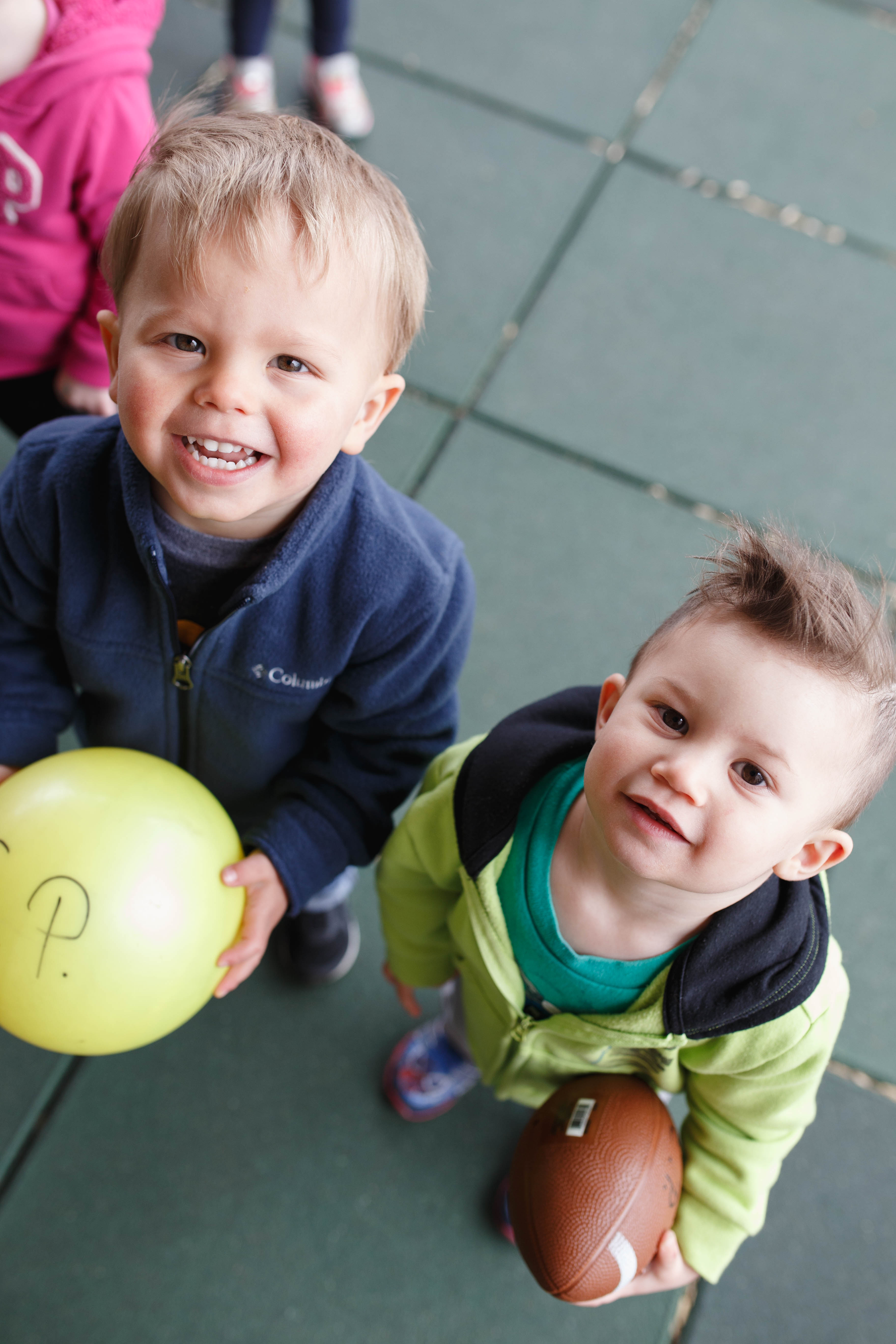 Two young boys standing on a green outdoor surface, each holding a football, looking up at the camera and smiling.