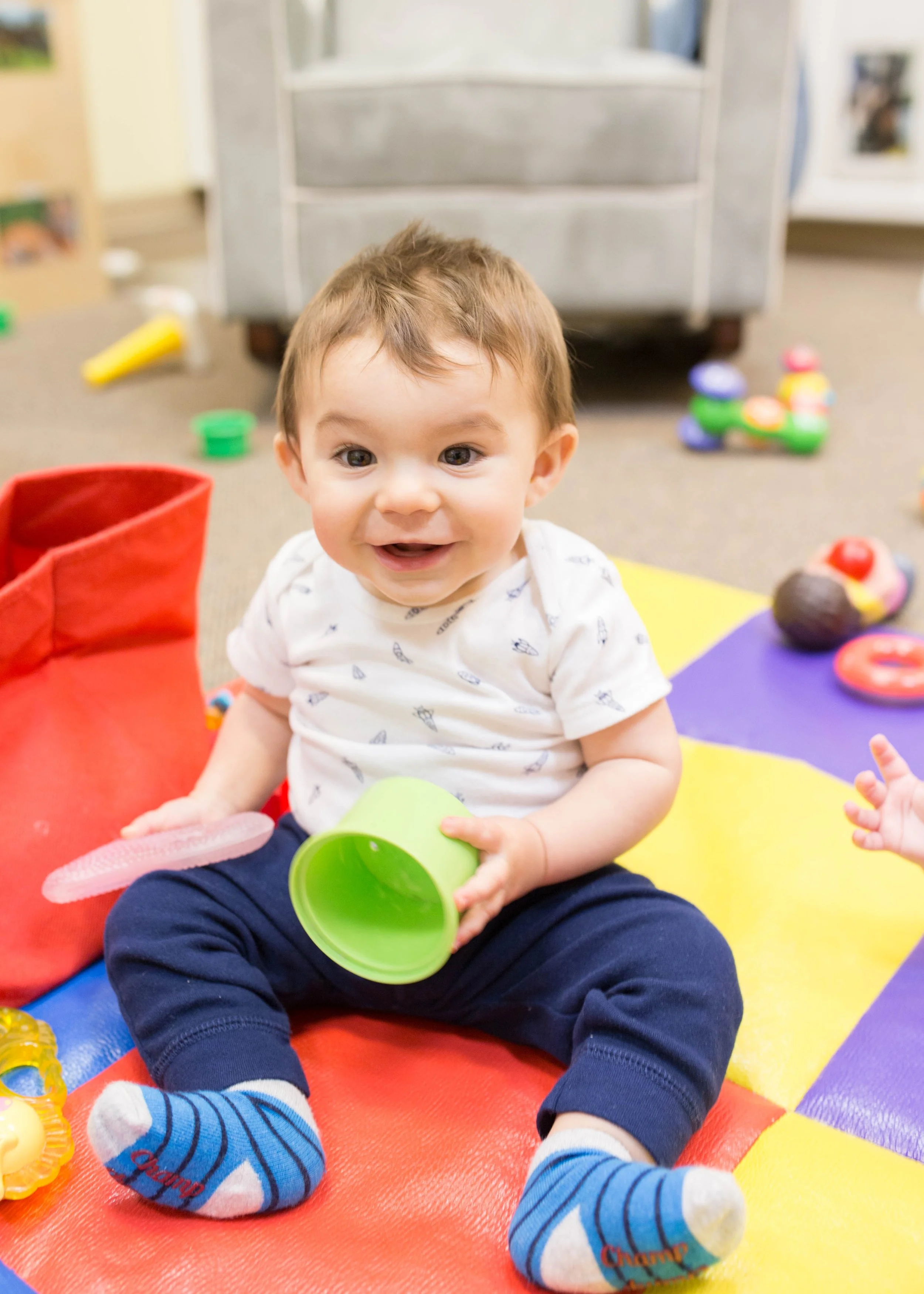 A baby boy sitting on a colorful foam mat playing with toys, smiling, surrounded by scattered toys and a toy storage unit in the background.